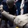 A worker wearing a gray balaclava face covering and work gloves is closely inspecting or working on a large metal pipe or tube resting on a sawhorse stand. Several other individuals in dark coveralls and work gloves are visible in the background, suggesting a group training or industrial work environment. The pipe shows visible surface marks, scratches, and what appears to be a small notch or defect near the worker's focus area.