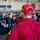 A person wearing a red uniform and a red cap embroidered with 'FREEDOM AT WORK' and 'XO' stands facing a group of personnel in a large indoor space, likely a naval vessel's hangar bay. The individual in the foreground is wearing what appears to be a championship-style belt across their shoulder, suggesting a recognition or award context. A crowd of uniformed personnel is gathered in the background, possibly for a ceremony or formation.