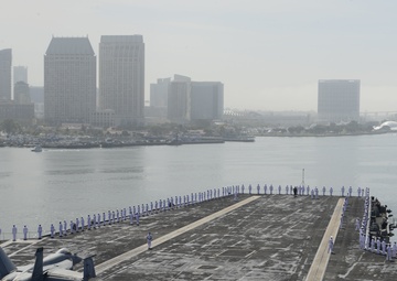 An aerial view of a U.S. Navy aircraft carrier flight deck with sailors in white dress uniforms manning the rails in a formal ceremonial formation. A single military jet aircraft, likely an F/A-18 Hornet, is parked on the flight deck. The carrier appears to be in a harbor with a city skyline visible in the background, consistent with San Diego, California.
