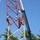 Radio communications tower erected at the 177th Fighter Wing of the New Jersey Air National Guard Two workers are climbing and performing maintenance on a red and white lattice communications or radio tower. They are equipped with safety harnesses and helmets, and a large crane boom is positioned adjacent to the tower, likely assisting with the work. Several buckets are suspended from ropes on the tower, presumably containing tools or materials.