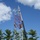 Radio communications tower erected at the 177th Fighter Wing of the New Jersey Air National Guard Workers are performing maintenance or construction on a red and white lattice communications tower, with multiple climbers visible at various heights on the structure. A crane with a blue boom is positioned alongside the tower, likely assisting with lifting equipment or materials. The scene includes support vehicles, safety fencing, and a small equipment building at the tower's base.