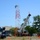 Radio communications tower erected at the 177th Fighter Wing of the New Jersey Air National Guard A construction or telecommunications site showing workers at height on a red and white lattice tower, assisted by a Link-Belt mobile crane. The scene depicts active tower assembly or maintenance work in what appears to be an industrial or semi-rural area. Multiple pieces of heavy equipment and safety barriers are visible on the ground.