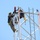 Radio communications tower erected at the 177th Fighter Wing of the New Jersey Air National Guard Two individuals in military camouflage uniforms are working at the top of a white metal lattice tower, likely performing maintenance or installation tasks. Both are equipped with full safety harnesses, helmets, and climbing gear, with orange safety ropes visible throughout. A red cylindrical device, possibly an aviation warning light with a solar panel, is mounted at the very top of the structure.