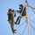 Radio communications tower erected at the 177th Fighter Wing of the New Jersey Air National Guard Two workers in military-style camouflage uniforms are performing maintenance or installation work at height on a white metal lattice tower or antenna structure. Both individuals are equipped with full safety harnesses, helmets, hearing protection, and climbing gear. They appear to be collaborating on a task involving equipment mounted on the tower's framework.