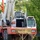 Radio communications tower erected at the 177th Fighter Wing of the New Jersey Air National Guard A large mobile crane truck marked with an 'OVERSIZE LOAD' banner is visible in an outdoor setting surrounded by green trees. At least one operator wearing a red hard hat is visible inside the cab. The vehicle appears to be part of an active construction or heavy lifting operation near a red metal structure.