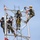 Radio communications tower erected at the 177th Fighter Wing of the New Jersey Air National Guard Four workers in military-style camouflage uniforms are performing maintenance or construction work at the top of a white metal lattice tower. All personnel are equipped with safety harnesses, lanyards, and hard hats, indicating adherence to fall protection protocols. Orange safety ropes are rigged throughout the structure, and the workers appear to be collaborating on a task at elevation against a clear blue sky.