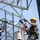 Radio communications tower erected at the 177th Fighter Wing of the New Jersey Air National Guard Two workers are performing high-elevation maintenance or construction work on a metal lattice tower or structure. Both are equipped with full safety harnesses, helmets, and are working with multiple colored ropes and rigging equipment. The image is taken from a low angle looking upward, emphasizing the height and complexity of the rigging setup.