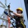 Radio communications tower erected at the 177th Fighter Wing of the New Jersey Air National Guard A worker is performing maintenance or installation work at height on a metal tower or mast structure. The individual is equipped with full fall-arrest safety harness, multiple carabiners, and colorful ropes, indicating adherence to working-at-height safety protocols. The image is taken from a low angle, emphasizing the elevated position of the worker against a clear blue sky.