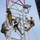 Radio communications tower erected at the 177th Fighter Wing of the New Jersey Air National Guard Four workers in military-style camouflage uniforms are climbing and working on a communications or utility tower structure. They are equipped with safety harnesses, hard hats, and tool buckets, suggesting a maintenance or installation operation at height. The tower appears to be a lattice-style steel structure with red and white coloring.