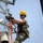 Radio communications tower erected at the 177th Fighter Wing of the New Jersey Air National Guard A worker in full safety gear is climbing or working on a white metal lattice tower or antenna structure. He is equipped with a fall arrest harness system, multiple ropes, and carabiners, indicating adherence to height safety protocols. The setting appears to be outdoors under a clear, hazy sky in warm conditions.