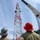 Radio communications tower erected at the 177th Fighter Wing of the New Jersey Air National Guard Two workers wearing hard hats observe a red and white communications or transmission tower under construction or maintenance. A Link-Belt crane is positioned to the right, assisting workers who are visible climbing or working near the top of the tower. The scene appears to be an active worksite with multiple personnel involved in the operation.