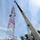 Radio communications tower erected at the 177th Fighter Wing of the New Jersey Air National Guard A Link-Belt crane is positioned next to a red and white lattice communications tower, with at least one worker visible near the upper section of the tower. The scene appears to depict tower construction, maintenance, or installation work taking place outdoors. The photo is taken from a low angle looking upward, emphasizing the height of both the crane boom and the tower against a partly cloudy sky.