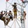 Radio communications tower erected at the 177th Fighter Wing of the New Jersey Air National Guard Four workers are performing maintenance or installation work on a communications or utility tower at significant height. The workers are equipped with full safety harnesses, helmets, and tool buckets, suggesting professional tower climbing operations. The team appears to be working in a coordinated manner on different sections of the white metal lattice tower.