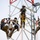 Radio communications tower erected at the 177th Fighter Wing of the New Jersey Air National Guard Four workers in military-style camouflage uniforms are performing maintenance or installation work on a communications or utility tower at significant height. All workers are equipped with safety harnesses, lanyards, and climbing gear, indicating adherence to fall protection protocols. The workers appear to be using hand tools and carrying equipment buckets while secured to the tower structure.