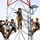 Radio communications tower erected at the 177th Fighter Wing of the New Jersey Air National Guard Four workers in military-style camouflage uniforms are performing maintenance or installation work on a communications or utility tower at significant height. All workers are equipped with safety harnesses, hard hats, and tool buckets, indicating adherence to fall protection protocols. The tower features red and white structural elements with what appears to be a solar panel or lighting fixture near the top.