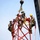 Radio communications tower erected at the 177th Fighter Wing of the New Jersey Air National Guard Three workers in military-style uniforms and safety harnesses are positioned at the top of a red lattice tower structure, connected to a crane hook via yellow lifting straps. The workers appear to be assembling or positioning the tower section, with a large crane visible on the right side of the frame. All personnel are equipped with safety gear including helmets and fall-arrest harnesses.