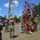 Radio communications tower erected at the 177th Fighter Wing of the New Jersey Air National Guard Military personnel in camouflage uniforms and hard hats are assembling or erecting a red metal tower structure at an outdoor work site. Several soldiers are actively working on the structure using a ladder, while others observe and supervise. A large crane with a red-and-white boom is visible in the background, suggesting a larger tower installation operation.