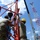 Radio communications tower erected at the 177th Fighter Wing of the New Jersey Air National Guard Two workers are assembling or rigging a red steel lattice structure, likely a tower or antenna base, using chains and lifting hardware. One worker wears a yellow hard hat and the other wears a patterned head covering, both dressed in military-style olive/tan uniforms. A red-and-white communications tower is visible in the background, along with a crane boom.