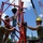 Radio communications tower erected at the 177th Fighter Wing of the New Jersey Air National Guard A group of workers in military-style uniforms and hard hats are assembling or rigging a large red steel structure using chains and a crane hook. The work appears to be taking place outdoors on a clear, sunny day with a large lattice crane visible in the background. The team is coordinating closely around the red metal frame structure.