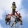 Radio communications tower erected at the 177th Fighter Wing of the New Jersey Air National Guard Several personnel in military-style camouflage uniforms are working at the top of a communications or antenna tower. They are equipped with safety harnesses, helmets, and climbing gear, suggesting a maintenance, installation, or training operation at significant height. The image is taken from a low angle looking upward against a partly cloudy blue sky.
