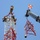 Radio communications tower erected at the 177th Fighter Wing of the New Jersey Air National Guard Workers in military-style or utility uniforms are climbing and working near the top of a red-and-white lattice communications tower. A crane is visible on the right side, suspending a separate tower section with a chain, suggesting active tower assembly or installation. An orange-and-white checkered aviation warning flag is attached to the crane boom.