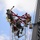 Radio communications tower erected at the 177th Fighter Wing of the New Jersey Air National Guard Three workers in military-style uniforms and safety harnesses are performing maintenance or construction work at the top of a communications or antenna tower. They are equipped with full fall-arrest harness systems, helmets, and tool bags. One worker has their arm raised, possibly signaling or reaching for equipment.