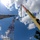 Radio communications tower erected at the 177th Fighter Wing of the New Jersey Air National Guard An upward-looking (worm's-eye view) photograph captures two cranes working in tandem near a red-and-white lattice communications tower. A white Link-Belt crane boom extends from the left while a yellow crane boom rises on the right, both appearing to be involved in tower construction or maintenance. The image is taken against a bright blue sky with scattered white clouds, with trees visible on the right edge.