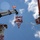 Radio communications tower erected at the 177th Fighter Wing of the New Jersey Air National Guard A low-angle upward shot captures a high-elevation construction or tower installation operation. Workers are positioned on a lattice tower structure on the left, on a suspended red-and-orange truss section being lifted by a crane in the center, and in an aerial work platform (cherry picker basket) on the right. The scene involves coordinated crane operations against a partly cloudy blue sky.