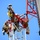 Radio communications tower erected at the 177th Fighter Wing of the New Jersey Air National Guard Several workers in military-style camouflage uniforms and safety helmets are performing maintenance or construction work at the top of a communications tower. The workers are equipped with full fall-arrest harness systems, lanyards, and safety gear while maneuvering on the red and white lattice structure. A large crane boom is visible on the right side of the frame, suggesting heavy lifting operations are underway.