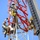 Radio communications tower erected at the 177th Fighter Wing of the New Jersey Air National Guard Three workers are performing high-altitude maintenance or construction work on a red and white lattice tower, likely a communications or antenna tower. All workers are equipped with full safety harnesses, helmets, and fall protection gear including lanyards and ropes. A large crane boom is visible on the right side of the frame, suggesting heavy equipment is being used in the operation.