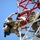 Radio communications tower erected at the 177th Fighter Wing of the New Jersey Air National Guard Two or more individuals in military/tactical camouflage uniforms are climbing or working on a red and white communications or radio tower. They are equipped with safety harnesses, gloves, and various tools attached to their gear. The photo is taken from a low angle looking upward, emphasizing the height and physical demands of the task.