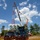 Radio communications tower erected at the 177th Fighter Wing of the New Jersey Air National Guard A large crane truck is actively working on a red and white lattice communications tower at an outdoor job site. The crane's boom is extended upward toward the top of the tower, with workers visible at elevation. The site is secured with orange safety fencing and surrounded by trees under a partly cloudy sky.