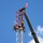 Radio communications tower erected at the 177th Fighter Wing of the New Jersey Air National Guard Several workers in safety harnesses and helmets are performing maintenance or rescue operations at the top of a red and white lattice communications tower. A large crane boom extends diagonally across the right side of the frame, positioned adjacent to the tower. The scene is photographed from a low angle against a bright blue sky with scattered clouds.