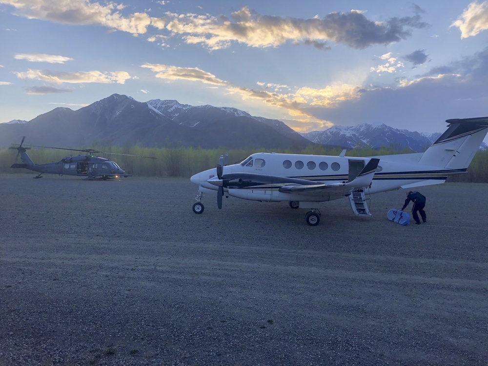 A twin-engine turboprop aircraft, likely a Beechcraft King Air, is parked on a gravel airstrip in a mountainous region during twilight or early dawn. A military-style helicopter, possibly a UH-60 Black Hawk, is visible in the background to the left. A person is crouched near the aircraft's airstair door, apparently loading or unloading equipment.