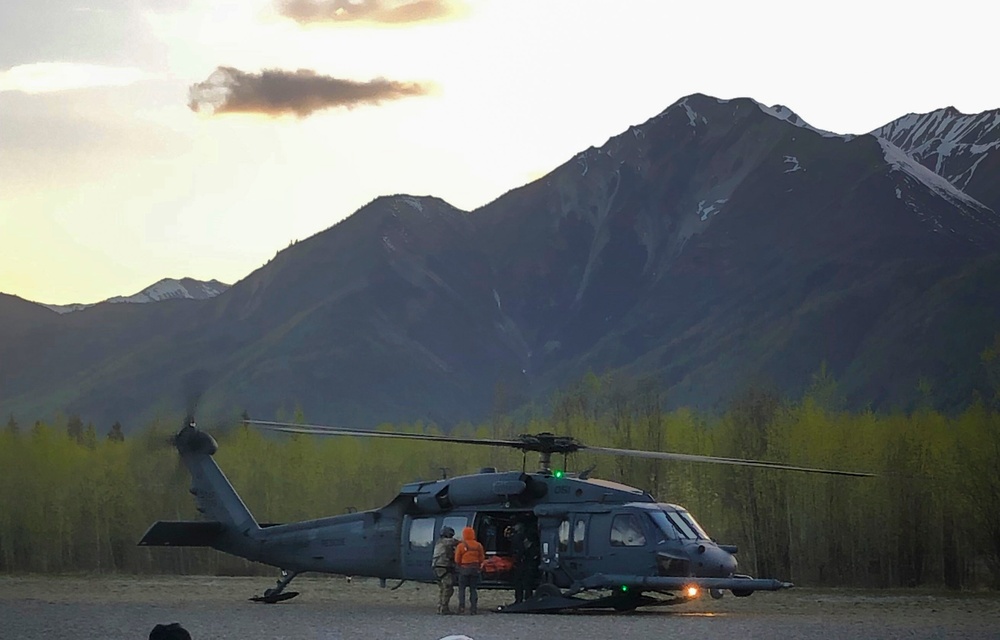 A military helicopter, consistent with an HH-60 Pave Hawk or similar Black Hawk variant, is grounded on a flat terrain area during twilight hours. Several personnel are visible near the open side door, with one individual wearing a bright orange jacket, suggesting a search and rescue or medevac operation. The scene is set against a dramatic backdrop of snow-capped mountains and spring foliage.