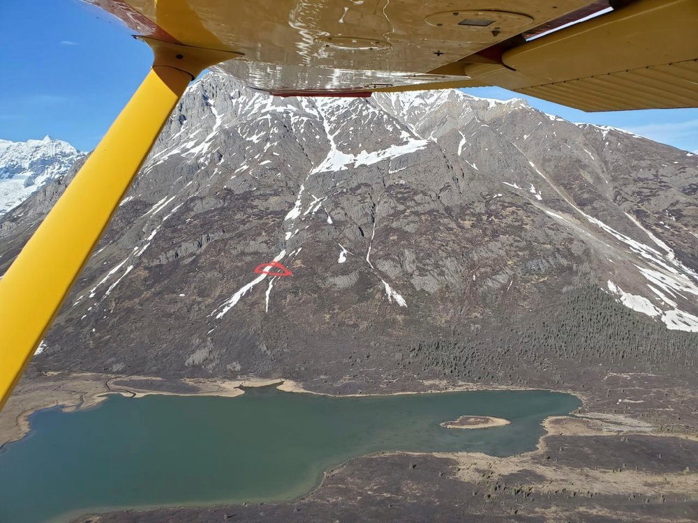 An aerial photograph taken from beneath the wing of a yellow small aircraft, showing a mountainous landscape with partial snow cover and a turquoise glacial lake below. A red circle or annotation is visible on the mountain face, likely marking a specific point of interest such as a geological feature or location. The scene appears to be in a remote wilderness area, possibly Alaska or the Canadian Rockies.