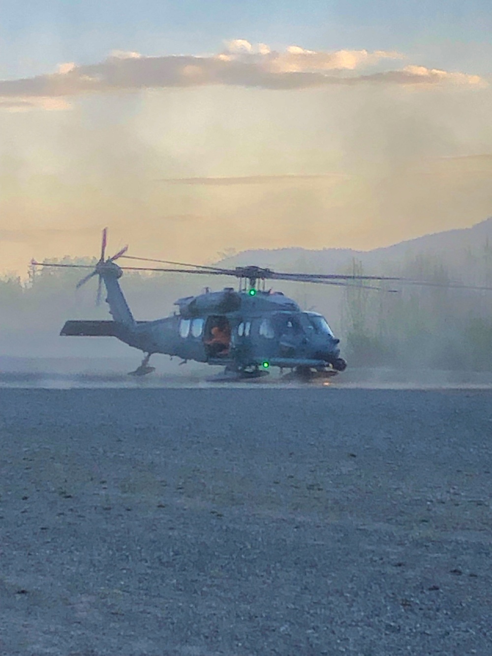 A military-style utility helicopter, consistent with a Sikorsky UH-60 Black Hawk or similar variant, is on the ground or hovering at very low altitude on a gravel surface. The scene appears to be at dusk or dawn with smoke or haze in the background, suggesting proximity to a wildfire or similar emergency operation. Rotor wash is visibly kicking up dust around the aircraft.