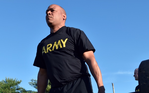 A U.S. Army soldier wearing a black 'ARMY' PT (physical training) shirt is performing a deadlift with a heavily loaded barbell outdoors. The low-angle shot emphasizes the weight and effort involved. Several other soldiers in matching Army PT uniforms are visible in the background, suggesting a group physical training session.