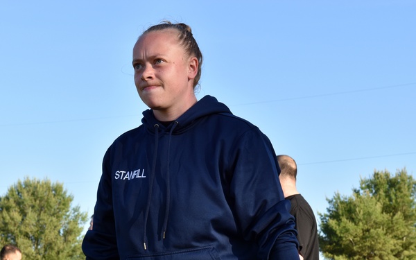 A person wearing a navy blue hoodie with 'STANFILL' printed on the chest is performing a deadlift or trap bar lift outdoors on a grass field. The individual is wearing shorts with 'ARMY' printed on them, suggesting a military fitness context. Several other people are visible in the background engaged in similar physical training activities.