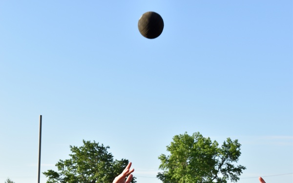 A person wearing a navy blue hoodie with the name 'STANFILL' is throwing or has just released a dark medicine ball upward during what appears to be a military fitness test or training exercise. In the background, a man in a black Army PT shirt holds a measuring pole, likely tracking the throw distance. The setting is an outdoor grassy field near military-style buildings.