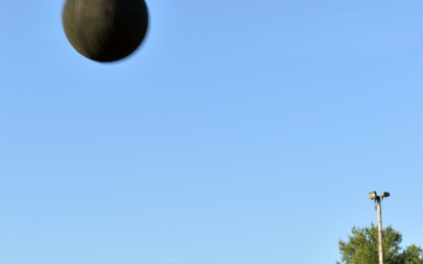 A young person wearing a black 'ARMY' t-shirt is throwing a medicine ball upward on an outdoor grass field. The action shot captures the moment just after release, with the dark ball visible in the upper portion of the frame against a clear blue sky. The setting appears to be a military or athletic training facility with orange cones marking the field.