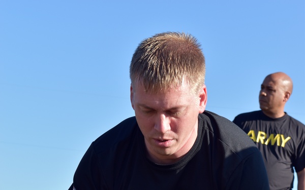 A U.S. Army soldier in black and gold PT (physical training) uniform is performing push-ups on a grass field outdoors. Two other soldiers in identical Army PT uniforms are visible in the background, one appearing to be resting and another standing. The setting appears to be a military physical fitness training session on a clear, sunny day.