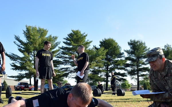 A group of U.S. Army soldiers are conducting a physical fitness test on a grass field. One soldier in PT (physical training) uniform is performing a prone exercise, likely a hand-release push-up, while a soldier in Army Combat Uniform (ACU) crouches nearby holding a clipboard to score or evaluate the performance. Additional soldiers in black Army PT uniforms observe or await their turn in the background.