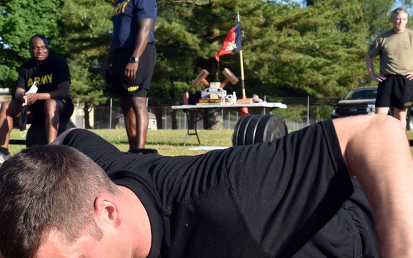 A soldier in black Army PT (Physical Training) uniform is performing a ground exercise, appearing to be in the low crawl or push-up position on a grass field. Several other soldiers and personnel in Army uniforms are visible in the background observing or resting. The setting appears to be an outdoor military physical fitness event or competition.