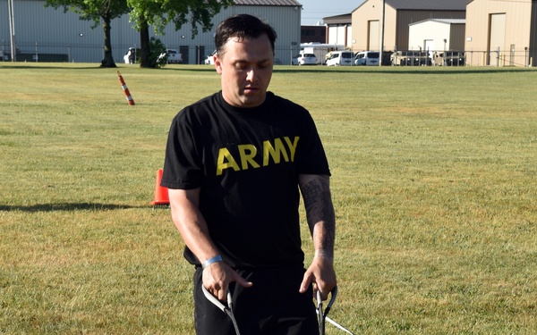 A man wearing a black U.S. Army physical training shirt and shorts is dragging a heavy weight sled across a grass field using two straps. The exercise appears to be part of a military fitness test or training event, likely the Army Combat Fitness Test (ACFT) Sprint-Drag-Carry event. The setting is an outdoor grassy area near military-style buildings.