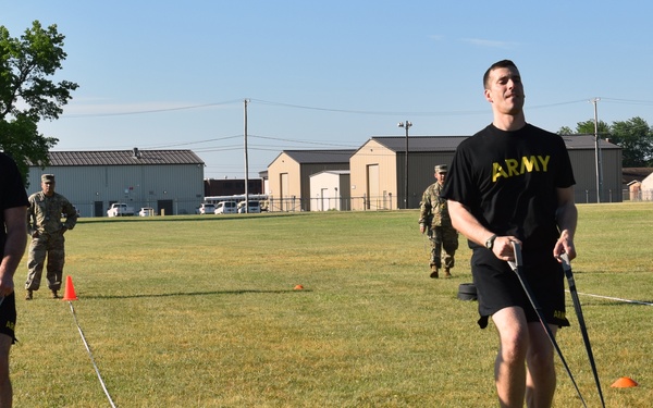 Two U.S. Army soldiers in black 'ARMY' PT shirts and shorts are performing a sled drag exercise on a grass field, pulling weighted sleds using cables. The exercise appears to be part of a physical fitness training event, with orange and yellow cones marking lanes and additional uniformed personnel observing in the background. The setting appears to be a military installation with warehouse-style buildings visible in the background.