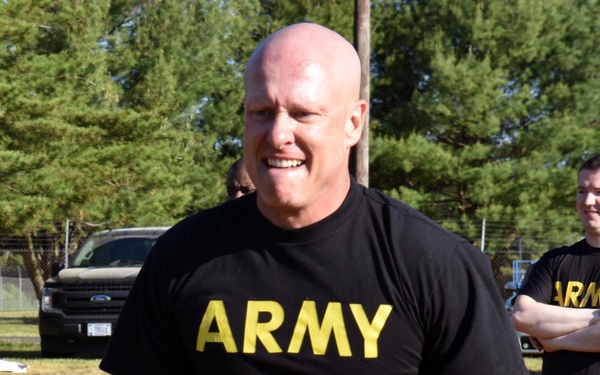 A bald male individual wearing black Army-branded athletic clothing is performing a farmer's carry exercise with two kettlebells on a grass field. The setting appears to be an outdoor military physical training event, with additional participants and equipment visible in the background. The subject is smiling, suggesting a positive or competitive atmosphere.