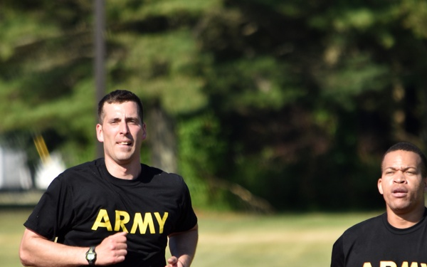 Two U.S. Army soldiers are running on a red athletic track, both wearing matching black Army PT (Physical Training) uniforms with yellow lettering. Additional runners are visible in the background, suggesting a group physical training event or fitness test. The setting appears to be a military installation with trees and a building visible in the background.