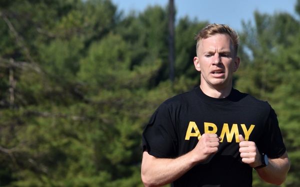 A young male soldier wearing U.S. Army physical training (PT) uniform is running on a red outdoor track. He appears to be exerting significant effort, with a focused expression and clenched fists. The setting is a well-maintained athletic facility surrounded by green trees under a clear sky.