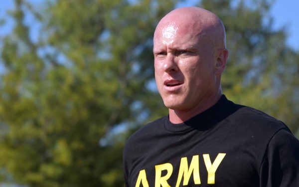 A bald male individual is running outdoors while wearing a black t-shirt with 'ARMY' printed in yellow letters. He appears to be sweating, suggesting sustained physical exertion. The background features green trees and a blue sky, indicating a daytime outdoor setting.